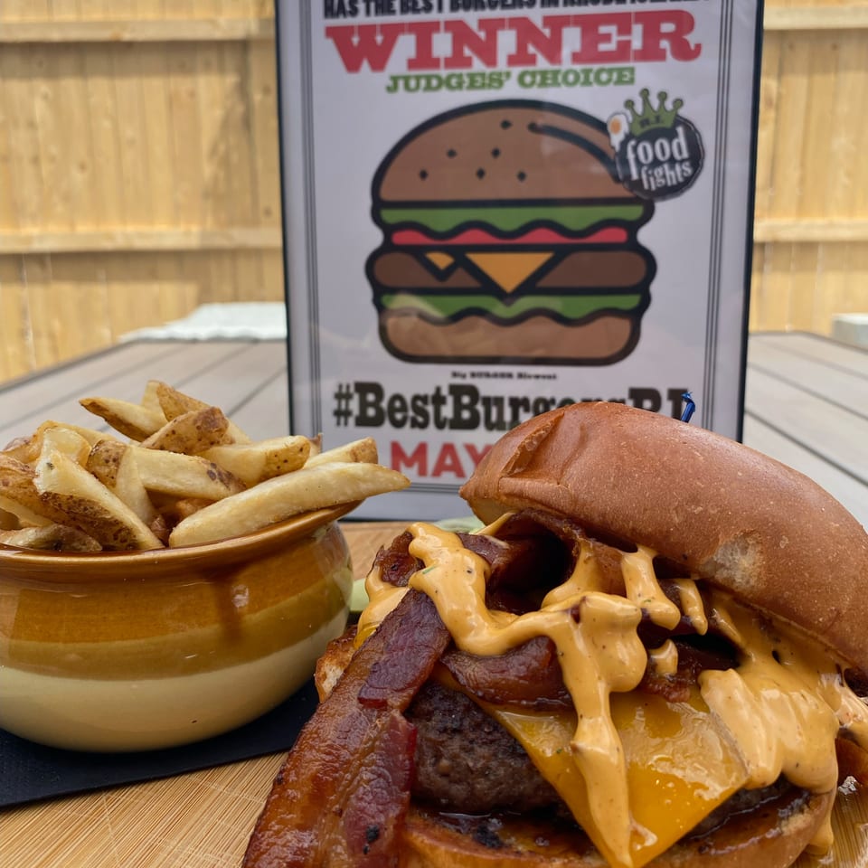 Bacon cheeseburger with creamy sauce and potato wedges on a wooden board, with a Rhode Island burger award sign in the background.
