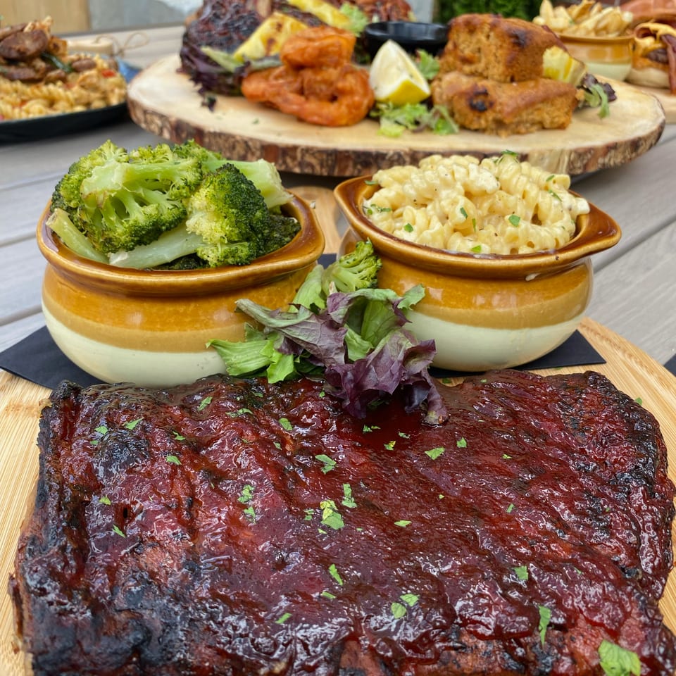 Barbecue feast with glazed ribs, grilled meats, falafel, fries, broccoli, creamy pasta, salad, and lemon wedges arranged on serving boards.