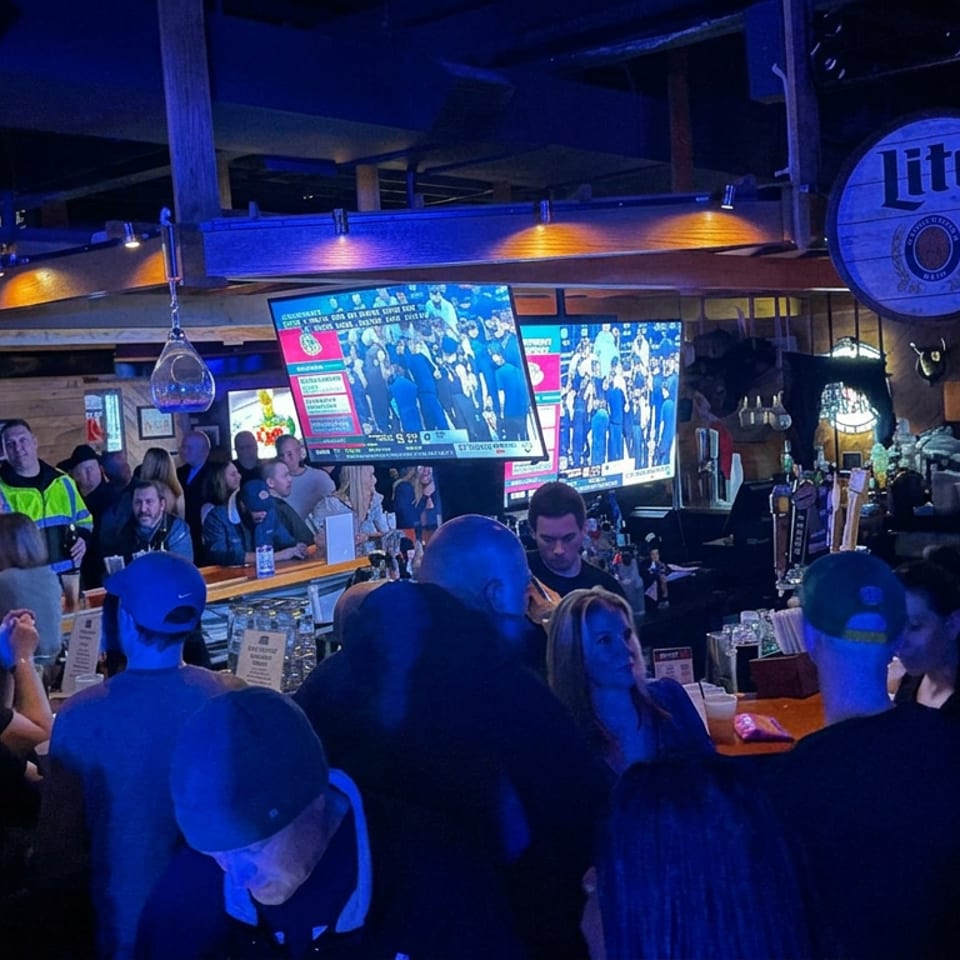 Busy sports bar with patrons gathered around the counter, multiple televisions showing a game, and Miller Lite branding overhead.