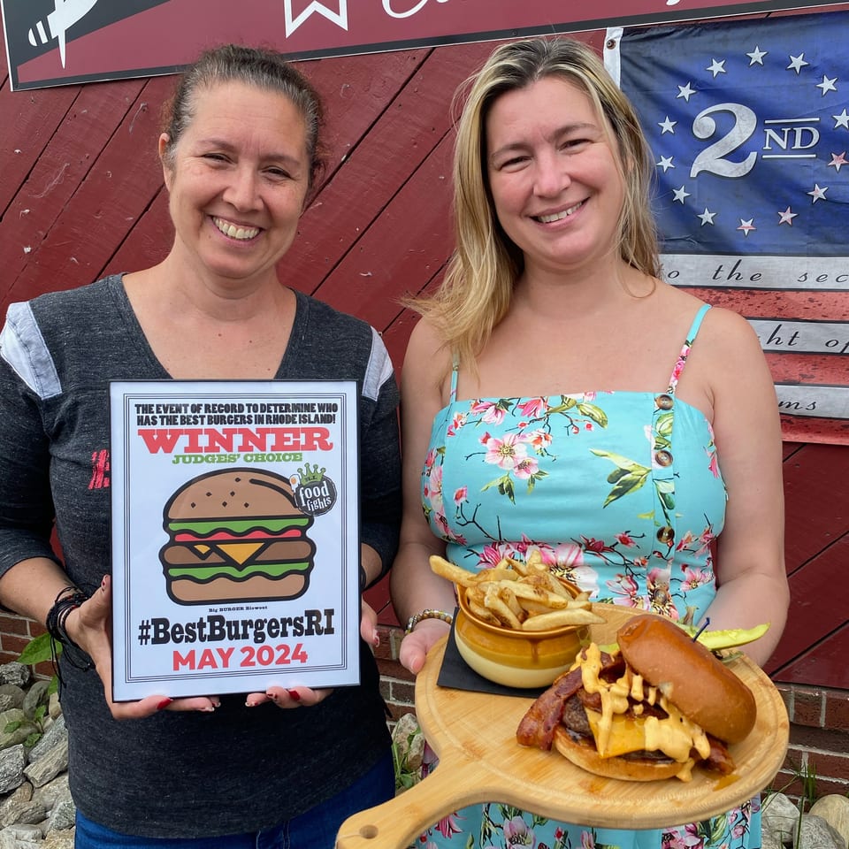 Two smiling women pose outside a restaurant with a best-burger award plaque, cheeseburger, fries, and dipping sauce.