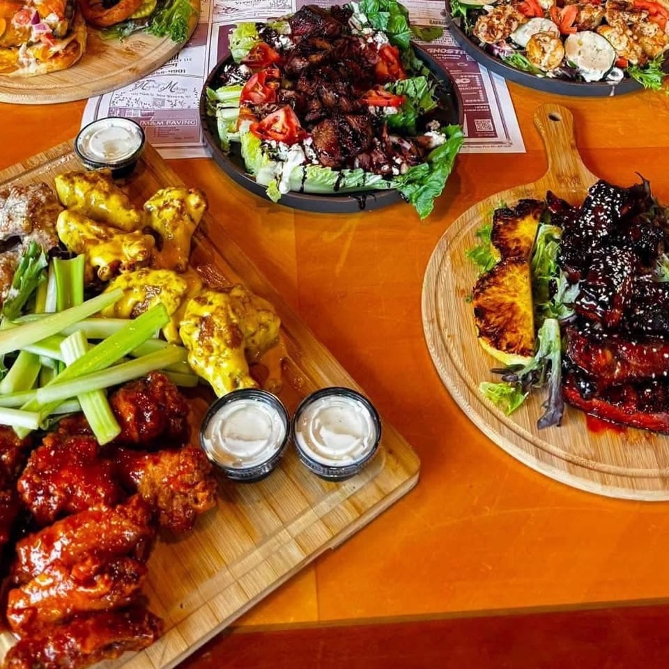 Assorted restaurant dishes, including sauced chicken wings, grilled meats, salads, vegetables, and dipping sauces, arranged on wooden boards.
