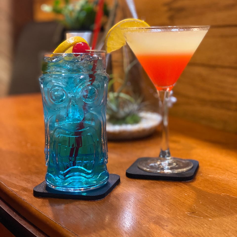 Blue iced cocktail with lime and cherry beside a red-orange martini, served on a wooden restaurant table.