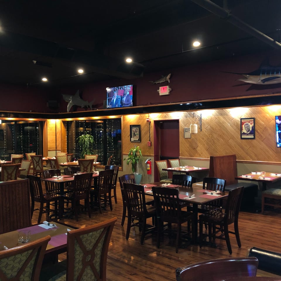 Spacious, warmly lit restaurant dining room with set tables, wood-paneled walls, mounted televisions, and fish decorations.